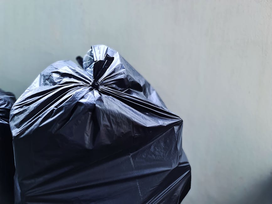 Close-up of a black rubbish bag made of shiny, crinkled plastic material, tied at the top with visible folds and creases, positioned in the foreground against a plain, light-colored wall. Additional rubbish bags are partially visible behind it, forming a stack or collection of waste waiting for collection or disposal. The scene appears to be on a driveway or pavement area, emphasizing the context of private waste handling and alternative waste collection services. Natural lighting highlights the reflective surface of the plastic bags, and the setting is neutral and professional, aligning with rubbish removal and waste disposal activities managed by waste disposal companies like Waste Disposals South Kensington.