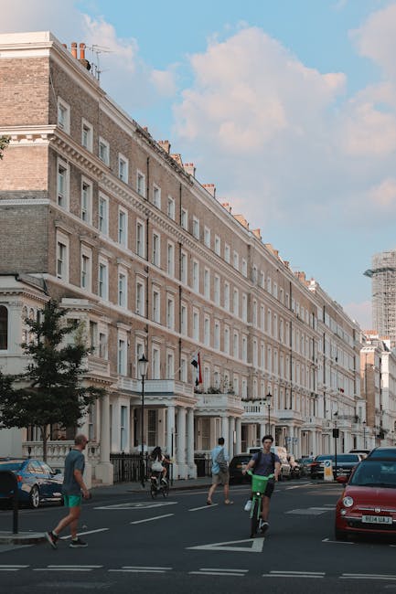 A row of multi-storey residential buildings with beige and white facades, decorative cornices, and large windows lining a busy urban street. Below, pedestrians walk along the pavement, and cyclists are riding on the road, which features marked lanes and parked cars on the curbside. The scene is set during daylight with a partly cloudy sky overhead, and a construction crane can be seen in the background. While the image showcases an active city environment, it highlights the importance of efficient rubbish removal and private waste handling services, such as those provided by Waste Disposals South Kensington, to maintain the cleanliness and order of bustling residential streets.