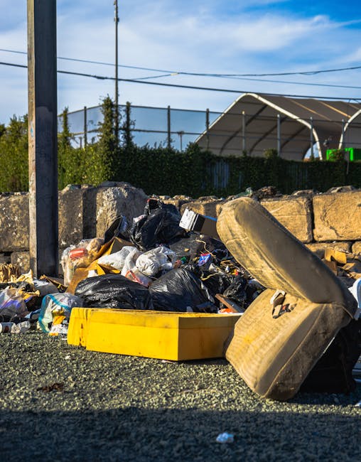 A pile of mixed household rubbish and waste materials, including black plastic trash bags, a large yellow plastic container, a worn and stained beige cushioned armchair, and various smaller debris, is seen on a gravel surface in an outdoor area. The waste is jumbled and appears to have been discarded illegally or left for collection. Behind the rubbish, there is a low stone wall made of roughly cut stones with cement between them, and above it, a dark green hedge borders the scene. In the background, a metal fence with vertical bars can be seen, along with some trees and a partially visible solar panel-covered structure. Overhead, power lines stretch across a bright blue sky with scattered clouds. The image illustrates a typical scenario requiring private rubbish removal or on-site clearance services, such as those offered by Waste Disposal South Kensington, to handle unauthorized or delayed waste disposal effectively.