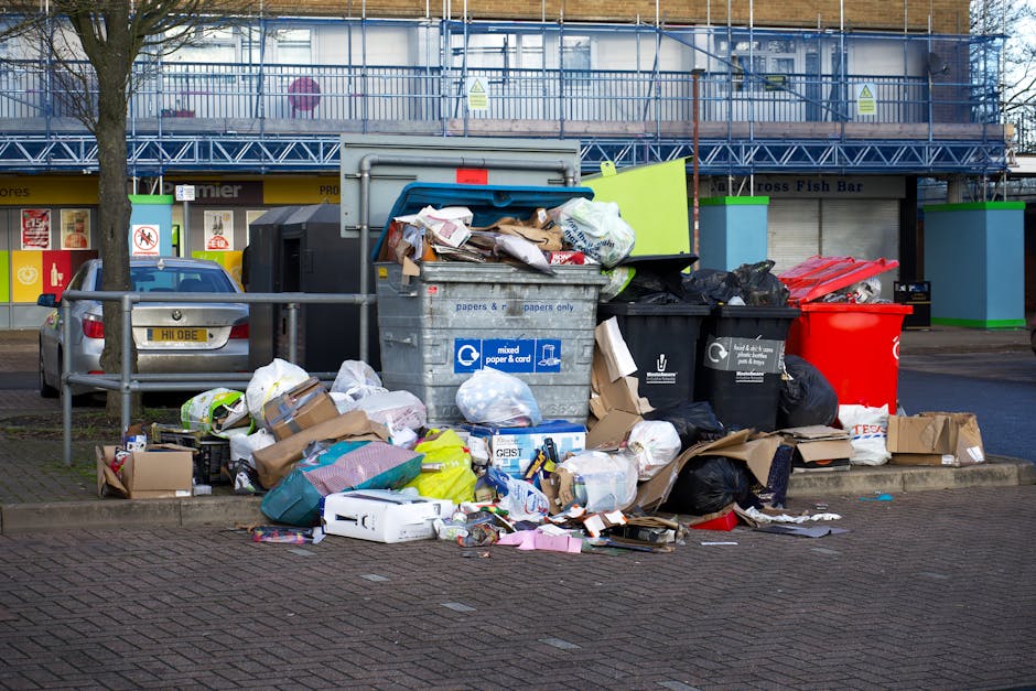 A large pile of mixed domestic waste and recycling bin bags is piled on a brick-paved sidewalk in an urban area, surrounding a grey communal waste container labeled for paper and cardboard. The waste consists of flattened cardboard boxes, plastic bags filled with trash, paper packaging, and some loose items such as cardboard tubes and plastic containers with various textures and finishes, predominantly in brown, black, white, and brightly coloured tones. Several black and red rubbish bags are seen around the container, some partially torn open, with loose rubbish spilling onto the surrounding pavement. The background features a building with a blue scaffolding structure and shopfronts, including signage and a display window, indicating a commercial or retail area. A parked car is visible behind a metal railing on the left, with a lantern and a leafless tree situated nearby, suggesting a typical urban scene. The overall atmosphere is neutral and observational, depicting a common scene of rubbish awaiting collection in a busy city environment, reflecting issues related to waste management and disposal.