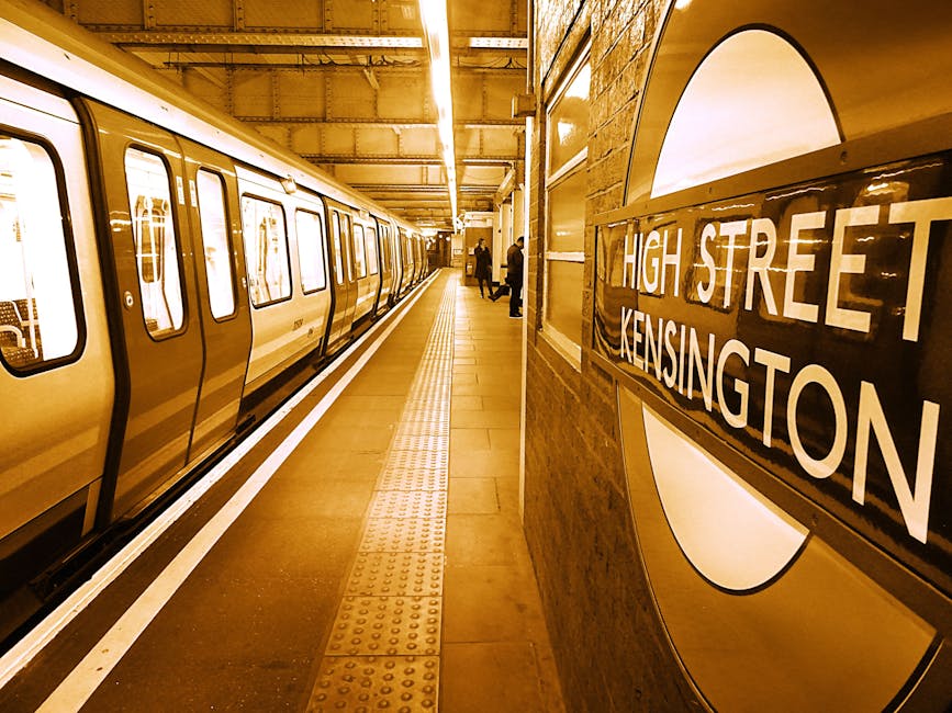 Inside a London Underground station platform at High Street Kensington, the image shows a portion of a mustard-yellow train with closed doors on the left side, parked alongside the platform. The platform surface is made of smooth, tan-colored tiles featuring tactile paving strips near the edge for safety. In the background, a few passengers are visible, some standing while others are walking along the platform. The station is illuminated with warm, ambient lighting, casting a gentle glow over the scene. On the right side, part of a large, circular sign is mounted on a brick wall, clearly displaying the station name 'HIGH STREET KENSINGTON' in white capital letters on a black rectangular background. The wall's texture is brick with a warm tone, complementing the overall lighting. This underground station setting, with its clearly visible signage, station environment, and waiting passengers, provides an authentic context related to urban transportation and the importance of maintenance or cleaning of public spaces, mirroring aspects of waste management and cleaning services, such as rubbish removal in busy public transit areas.