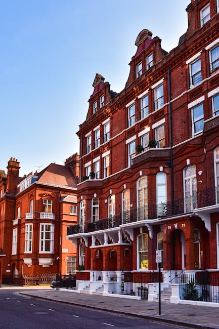 The image depicts a row of historic red-brick residential buildings along a city street in South Kensington, featuring multiple floors with large sash windows, some with small balconies or iron railings. The buildings have decorative architectural details such as ornate cornices, arched windows, and brickwork patterns, with the upper floors showing attic windows set within dormer structures. In the foreground, the pavement is visible alongside a section of the street, with a street sign, a black waste bin, and metal railings lining the entrance steps of one building. The sky above is clear and blue, suggesting a bright and sunny day. The scene illustrates an area where private waste disposal or rubbish collection could occur, with the setting typical of outskirts of upscale residential districts, aligned with independent rubbish removal services operating in South Kensington. The detailed features and materials, such as the textured brickwork and contrasting white trim, are accurately captured, providing a comprehensive visual description suitable for accessibility purposes and natural SEO integration related to rubbish removal services.
