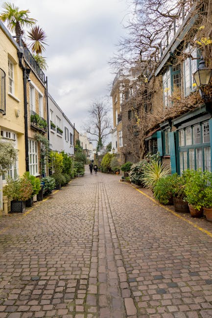 A narrow cobblestone street lined with residential buildings on both sides, featuring a mix of modern and older architectural styles. The left side has cream-colored buildings with large windows framed in white, some with window boxes of greenery, while the right side shows darker brick structures with blue painted wooden extensions and glass emporiums. Potted plants with lush green foliage and palm-like trees are positioned along the pavement, adding a touch of nature to the scene. In the background, two people are walking along the street amidst overhanging trees with bare branches, indicating a season of late autumn or early winter. The sky is cloudy, casting diffuse natural light over the area, which appears quiet and tidy. This scene exemplifies an urban environment where private, non-council waste collection could be coordinated, reflecting the context of independent rubbish removal and waste management services that operate in residential neighbourhoods.