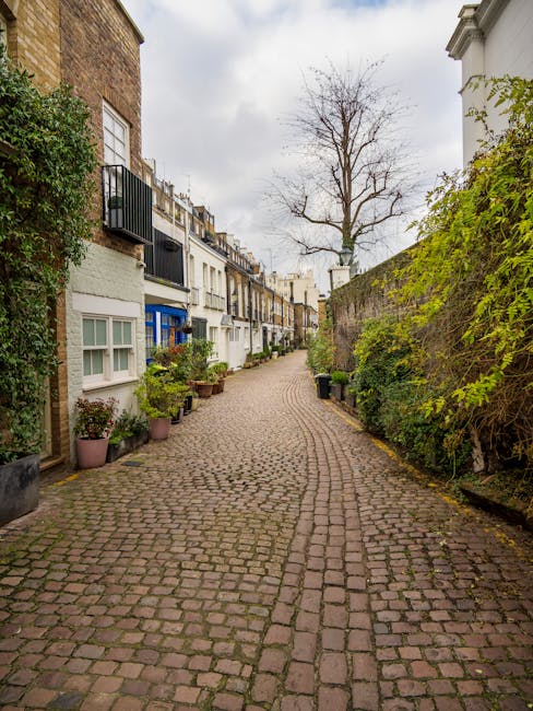 A narrow cobblestone street in a residential area, bordered by a row of multi-story terraced houses with small front gardens and potted plants. The buildings display various colours, including white, beige, and light blue, with some featuring black iron railings and window frames. On the right side, a tall, leafless tree stands behind a high, weathered brick wall covered partially with green foliage. The street appears quiet, with no vehicles or pedestrians visible, and overcast grey skies cast diffuse light over the scene. This setting exemplifies an urban environment where private waste collection or on-site clearance might be undertaken, with no visible rubbish or waste materials present, supporting local rubbish removal services that focus on discreet, independent collection within residential streets like this one.