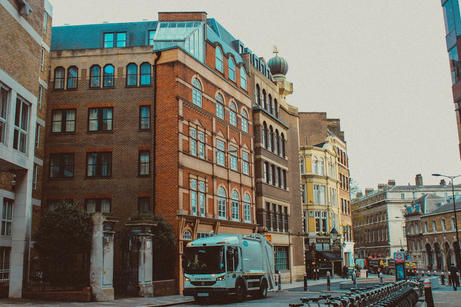 The image depicts a row of light-colored, multi-storey residential buildings with classic architectural detailing, including decorative cornices and large windows, situated along a city street with a clear blue sky and a few scattered clouds overhead. In the foreground, pedestrians are crossing the street, with some walking, a cyclist riding a green bicycle, and others waiting or standing nearby. Several parked cars line the roadside, including a red vehicle on the right and a darker vehicle further down. The street appears busy but orderly, with some trees providing minimal greenery on the sidewalk. Visible in the background, a construction site with a high-rise building under development can be seen, along with a lamppost and flagpoles attached to the buildings. The scene conveys a typical urban environment, with a subtle connection to waste disposal and rubbish removal services, supported by the neutral setting and the context of private waste handling possibly provided by local waste disposal companies like Waste Disposals South Kensington, visible through their reference to alternative collection services in the area.