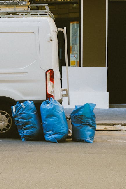 A white commercial van parked on a street in front of a building with large glass windows and a white exterior wall, with three blue rubbish bags placed on the pavement beside the vehicle. The bags are made of plastic, slightly crumpled, and filled with unidentified waste materials. The rear side of the van features a vertical tail light and a partially visible wheel with a multi-spoke alloy rim. The scene appears to be part of a private waste disposal or rubbish removal service, with the bags positioned for collection, reflecting an alternative to regular municipal rubbish collection methods. The environment is urban, illuminated by natural daylight, and the ground surface is paved asphalt, showing a typical setting for independent rubbish clearance or on-site waste handling, as often managed by waste disposal specialists like those at wastedisposalsouthkensington.co.uk.
